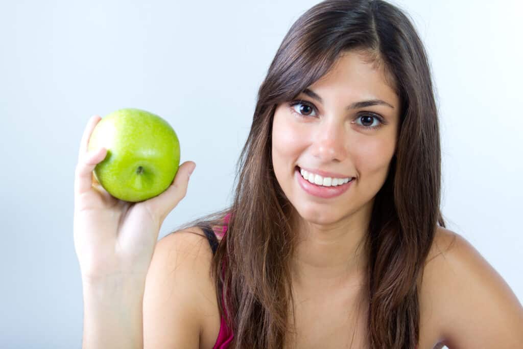 Mujer sosteniendo una manzana verde, es muestra de una sonrisa saludable y estética, sinónimo de una buena salud dental